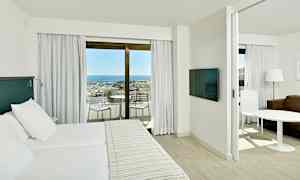 Et hotelværelseinteriør med en dobbeltseng, et separat siddeområde og en altan med udsigt over havet og en marina. (AI-genereret tekst)