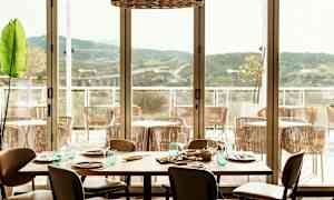 En restaurant med et dækket bord og udsigt over en terrasse med stole og bjerge i det fjerne. (AI-genereret tekst)
