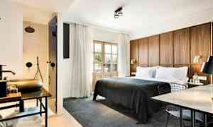 Et hotelværelse med en stor seng dækket med mørkegråt sengetæppe, en træpanel som sengegavl, et arbejdsområde med bord og stol, samt et synligt badeværelse med bruser bag en glasdør. (AI-genereret tekst)