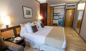 Et hotelværelse med en stor seng, puder, natborde og en lille køkkensektion med skabe. (AI-genereret tekst)