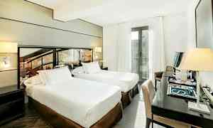 Et hotelværelse med to enkeltsenge, et skrivebord med en stol og en altan med hvide gardiner. (AI-genereret tekst)