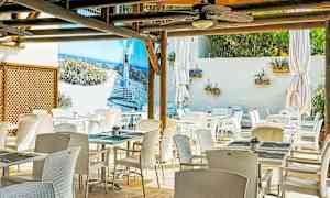 En udendørs restaurant med hvide stole og borde. En væg har et stort billede af en strandpromenade. (AI-genereret tekst)