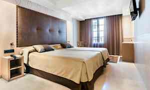 Et hotelværelse med en stor seng, brun sengegavl og beige sengetæppe. Et tv er monteret på væggen, og vinduet er dækket med mørke gardiner. (AI-genereret tekst)