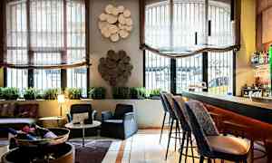 Et indendørsbillede, der viser en lounge eller barområde med stole, et bord, en bardisk og dekorative elementer på væggene. (AI-genereret tekst)