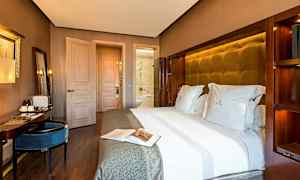 Et hotelværelse med en stor seng redt med hvidt sengetøj, et skrivebord med siddeplads og en dør, der fører til badeværelset. (AI-genereret tekst)