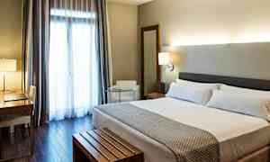 Et hotelværelse med en dobbeltseng, et skrivebord med stol, en lænestol og et stort vindue med gardiner. (AI-genereret tekst)