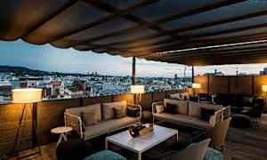 En terrasse med tag og udsigt over en bys skyline. Terrassen er møbleret med sofaer og borde. (AI-genereret tekst)