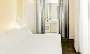 Et hotelværelse med en hvid seng og et moderne badeværelse synligt gennem lodrette sorte paneler. (AI-genereret tekst)