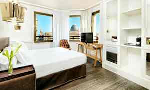 Et hotelværelse med en dobbeltseng, mørkt trægulv og et skrivebord med TV. Værelset har store vinduer med udsigt over byen. (AI-genereret tekst)