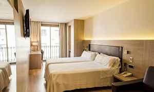 Et hotelværelse med to enkeltsenge, et skrivebord og en stol foran et stort vindue med altangelænder. Et TV er monteret på væggen. (AI-genereret tekst)