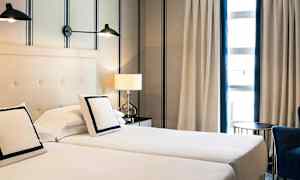 To enkeltsenge står side om side med puder og hvidt sengetøj. Bag sengene er en lys væg med stribemønster. Ved siden af sengen er et bord med en bordlampe, og et vindue med gardiner ses i baggrunden. (AI-genereret tekst)