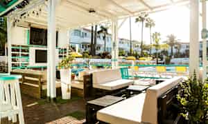 En udendørs lounge under en hvid pergola med møbler og planter, med udsigt over en pool og omkringliggende bygninger. (AI-genereret tekst)