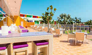 En udendørsterrasse med borde og stole, omgivet af palmer og farverige bygninger. (AI-genereret tekst)