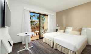 Et moderne hotelværelse med to enkeltsenge, et lille bord og et vægmonteret tv. Værelset har lyse farver og et vindue, der fører ud til en balkon med udsigt til palmer. (AI-genereret tekst)