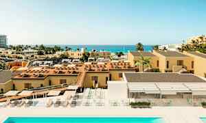 Et billede af et ferieområde med gule bygninger, en swimmingpool, liggestole og udsigt over havet i det fjerne. (AI-genereret tekst)