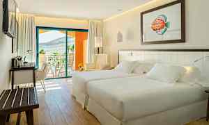 Et lyst og moderne hotelværelse med to senge, et siddeområde og et skrivebord. En balkon med udsigt over landskabet ses i baggrunden. (AI-genereret tekst)