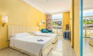 Et hotelværelse med to enkeltsenge, en blå sofa og store vinduer med udsigt udenfor. (AI-genereret tekst)