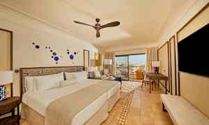 Et hotelværelse med en dobbeltseng, siddepladser, et skrivebord og et tv. Værelset har en moderne indretning med blå dekorationer på væggen og et vindue, der fører ud til en balkon med udsigt. (AI-genereret tekst)