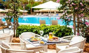 Et bord med mad og drikke står på en udendørs terrasse nær en pool omgivet af blomstrende planter og parasoller. (AI-genereret tekst)