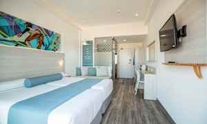 Et moderne hotelværelse med en dobbeltseng dekoreret med blå tekstiler, en siddeplads med puder, et skrivebord og et vægmonteret fjernsyn. (AI-genereret tekst)