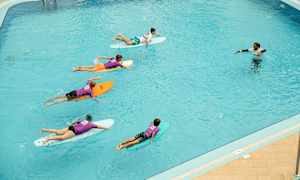 En gruppe personer ligger på surfbrætter i en pool, og en instruktør står i vandet. (AI-genereret tekst)