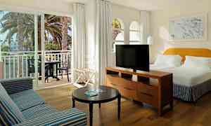 Billede af et hotelværelse med en stor seng, en sofa, et bord og et TV. En balkon med bord og stole ses gennem skydedøre. (AI-genereret tekst)