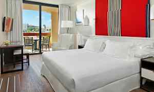 Et hotelværelse med en stor seng, en lænestol, et bord med stole og en altan med udsigt over en have. (AI-genereret tekst)