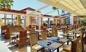 En udendørs restaurant med siddepladser bestående af borde med kurvestole. Solsejl dækker taget, og palmer kan ses i baggrunden. (AI-genereret tekst)