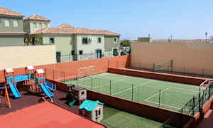 Billedet viser en udendørs legeplads med rutchebane og legehuse samt en grøn indhegnet sportsbane til fodbold og volleyball. I baggrunden ses boliger. (AI-genereret tekst)