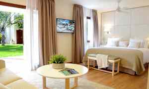 Et lyst hotelværelse med en stor seng, vægmonteret TV og en siddegruppe foran et rundt bord. Store gardiner med udsigt til en have. (AI-genereret tekst)