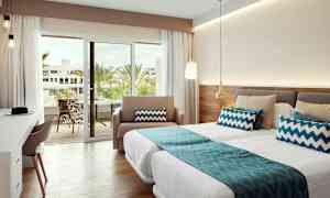 Et veloplyst hotelværelse med senge, blå dekorative puder og en glassdør, der fører til en balkon med udsigt over palmer. (AI-genereret tekst)