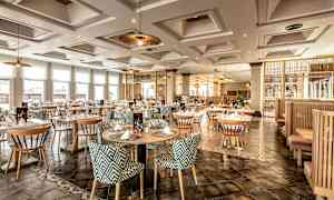 Et elegant restaurantområde med runde borde og mønstrede stole, belyst af loftslamper og naturligt lys gennem store vinduer. (AI-genereret tekst)