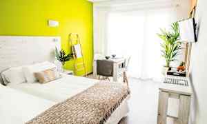 Et lyst og moderne soveværelse med grøn accentvæg, seng med hvidt og beige sengetøj samt bord og stole tæt på et stort vindue. (AI-genereret tekst)