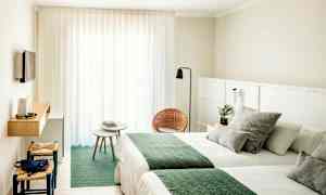 Et hotelværelse med lyse vægge, to senge med grønne tæpper, en lille siddegruppe med stole og bord samt et vægmonteret TV. (AI-genereret tekst)