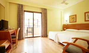 Et lyst hotelværelse med to senge, skrivebord, stol og balkon med glasdøre. (AI-genereret tekst)