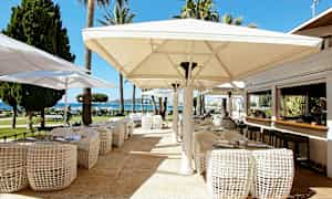 Udendørs serveringsområde med borde, stole og store parasoller, med udsigt mod have og hav. (AI-genereret tekst)