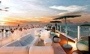 Tagterrasse med udsigt over havet ved solnedgang, med moderne møblering inklusive sofaer, parasoller og borde. (AI-genereret tekst)