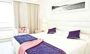 Et lyst hotelværelse med en dobbeltseng og indretning i lilla og hvidt. (AI-genereret tekst)