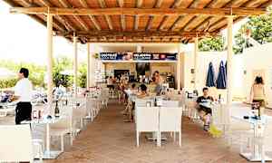 En udendørs siddeområde ved en bar med trætag, borde og stole. Folk sidder ved bordene og bestiller fra baren. (AI-genereret tekst)