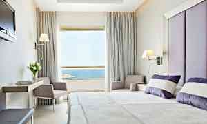 Et hotelværelse med moderne indretning. Sengen er redt med hvide og lilla puder, og der er et stort vindue med udsigt over havet. (AI-genereret tekst)