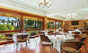 En elegant restaurant med dækkede borde og fletstole. Store vinduer giver udsigt til en have. (AI-genereret tekst)