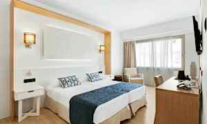 Et hotelværelse med to senge med blå sengetæppe og mønstrede puder, skrivebord, stol og en lænestol ved et vindue. (AI-genereret tekst)