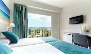 Et hotelværelse med to enkeltsenge, et maleri med strandmotiv på væggen, store vinduer med udsigt over landskabet og et TV på væggen. (AI-genereret tekst)