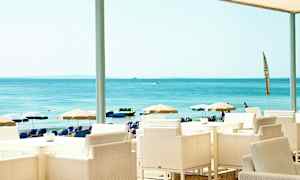 Udsigt fra en strandrestaurant med hvide stole og borde, i baggrunden ses parasoller og havet. (AI-genereret tekst)