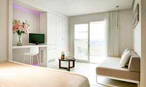 Et lyst hotelværelse med en seng, en sofa, et bord og en balkon med udsigt over havet. (AI-genereret tekst)