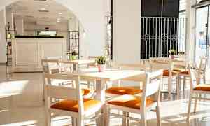 Lyse træmøbler med orange sædehynde i et solrigt rum med store vinduer. (AI-genereret tekst)