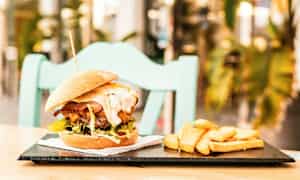 En burger med tilbehør af pommes frites på en tallerken, placeret på et bord med en stol i baggrunden. (AI-genereret tekst)