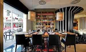 Interiør af en moderne restaurant med træborde og sorte stole, omgivet af stribede vægdekorationer og store vinduer. (AI-genereret tekst)