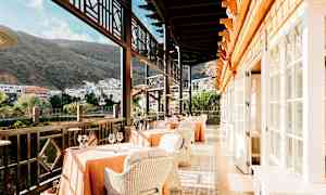 Restaurant med borde og stole på en terrasse med udsigt over bygninger og bjerge. (AI-genereret tekst)