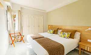 Et hotelværelse med to enkeltsenge, dekorative puder og et brunt sengetæppe samt en garderobe og en stol ved vinduet. (AI-genereret tekst)